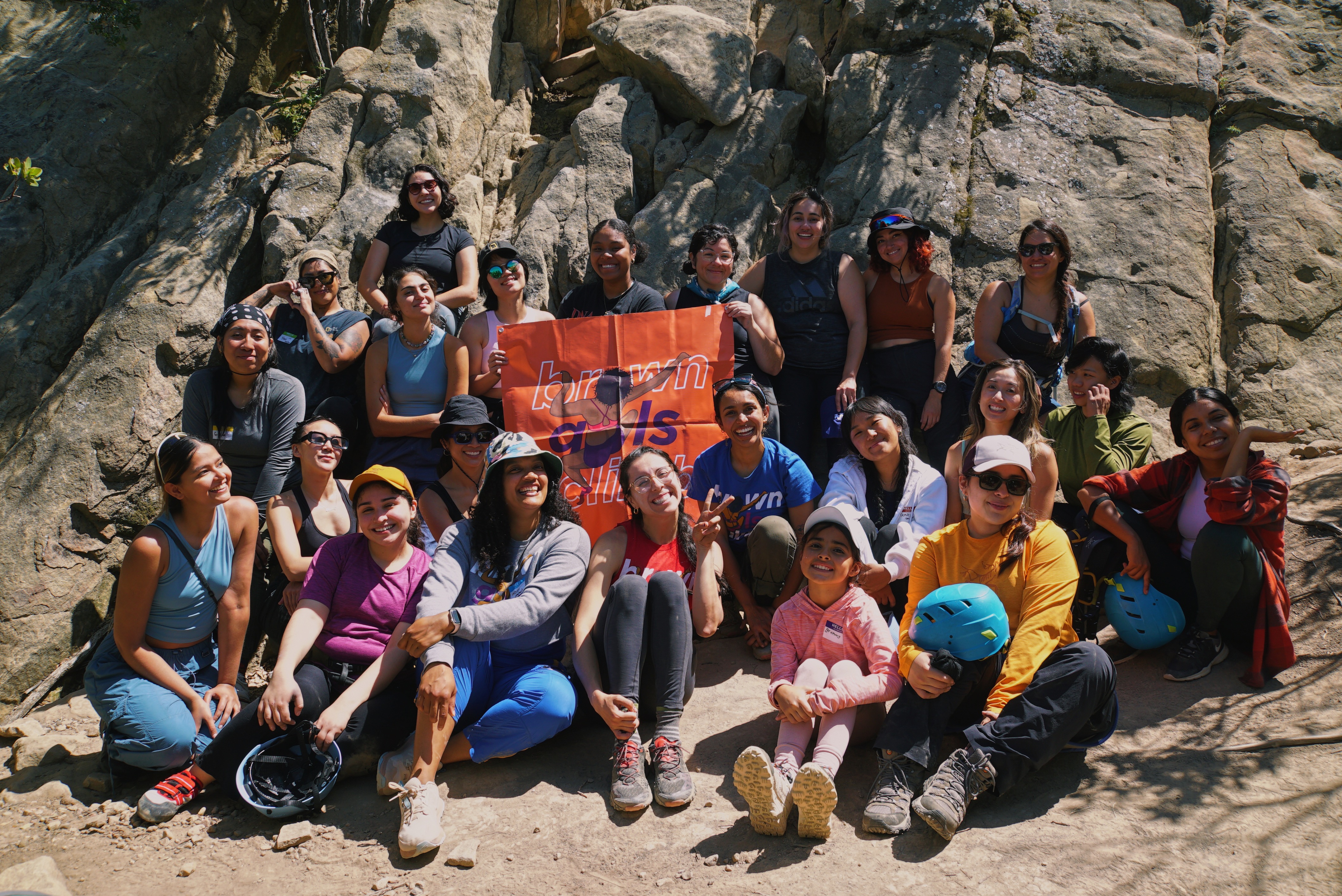 A group of people sit around a Brown Girls Climb flag next to a cliff